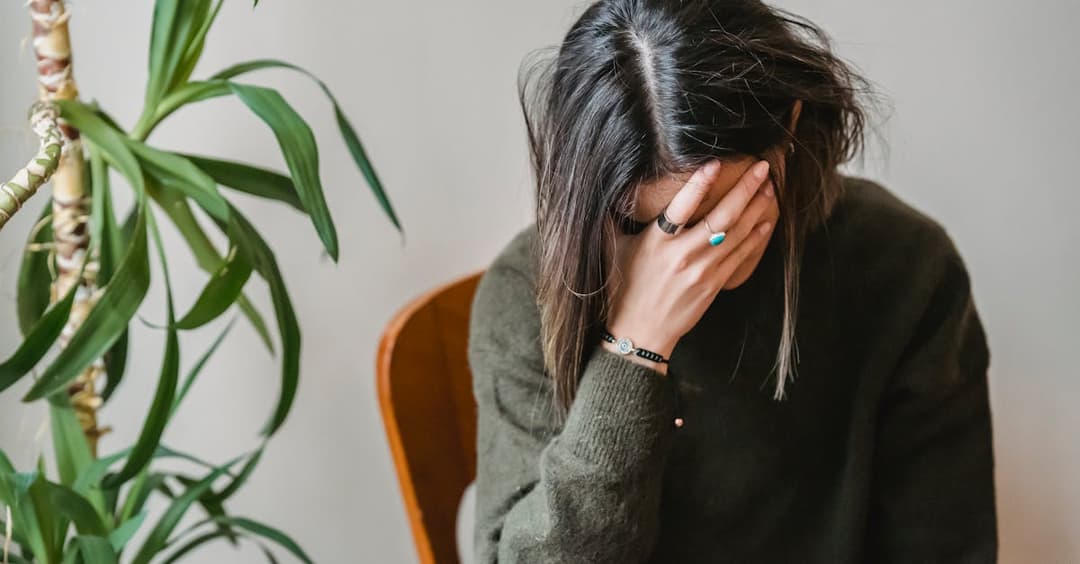 Unrecognizable young upset female millennial with dark hair in stylish sweater covering face with hand while siting on chair at home