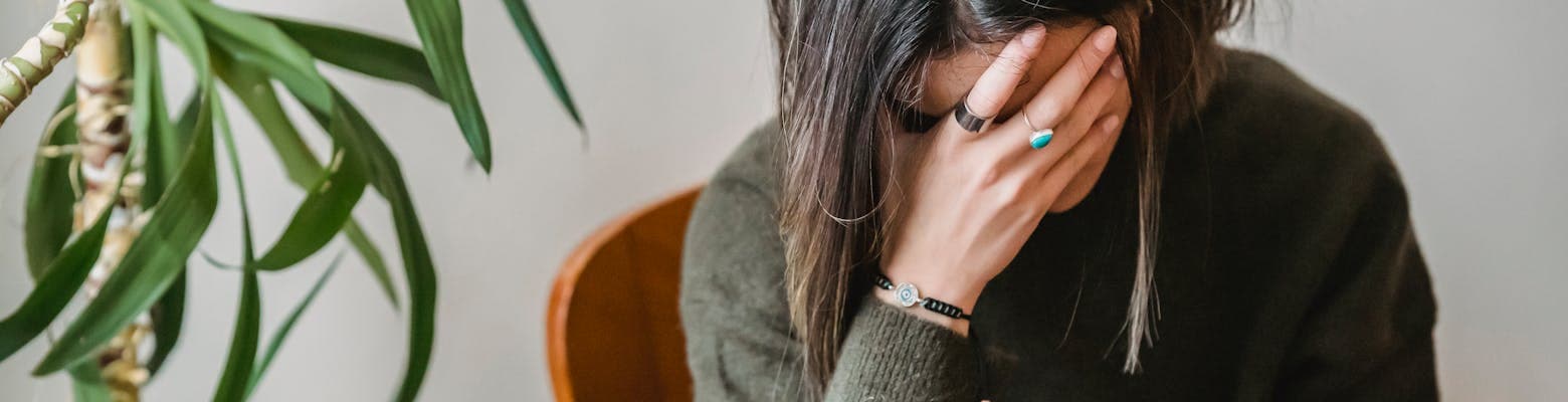 Unrecognizable young upset female millennial with dark hair in stylish sweater covering face with hand while siting on chair at home